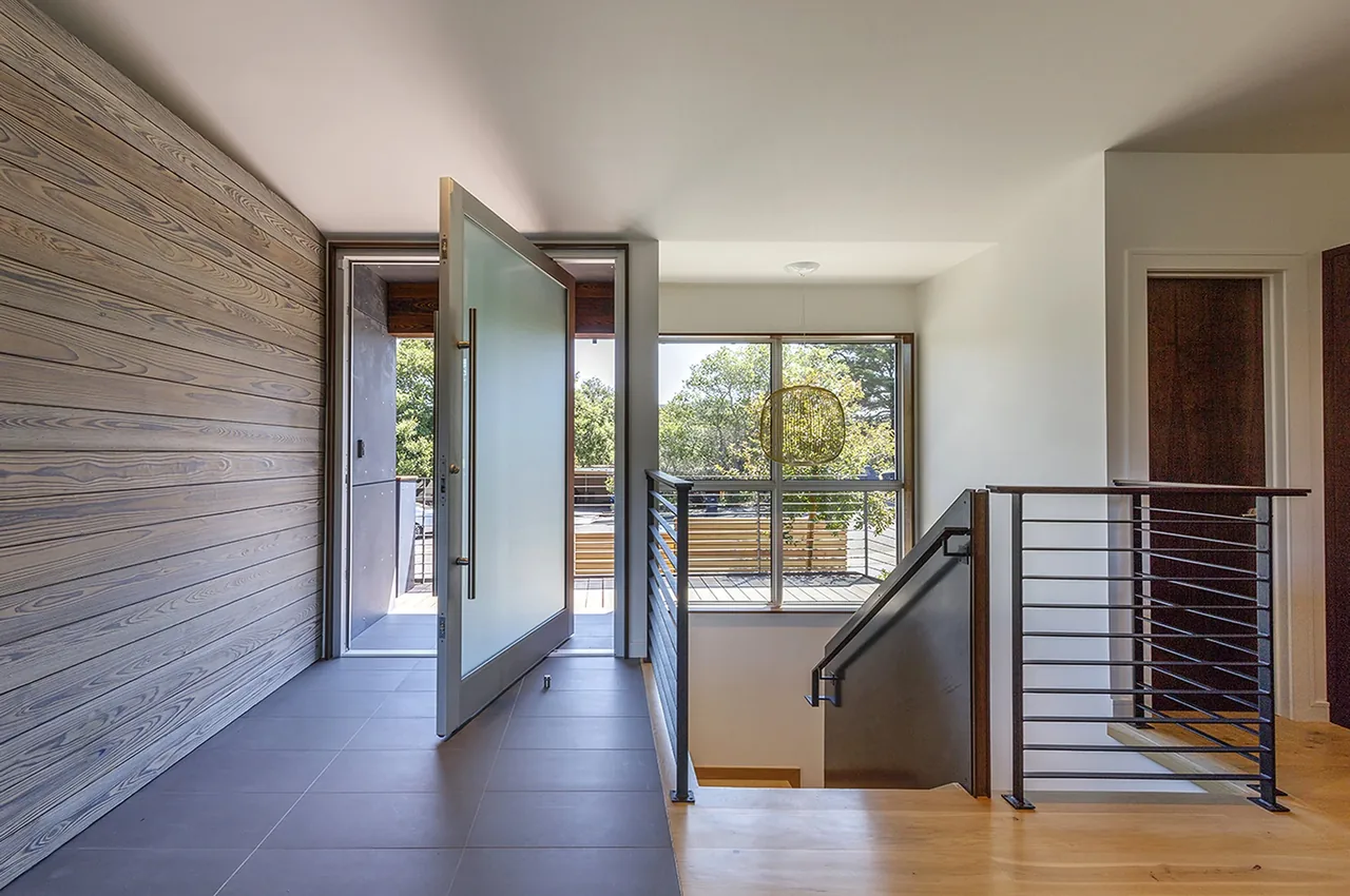Open glass entry door reveals balcony with horizontal metal railing and wood exterior siding at Shou Sugi Ban House.