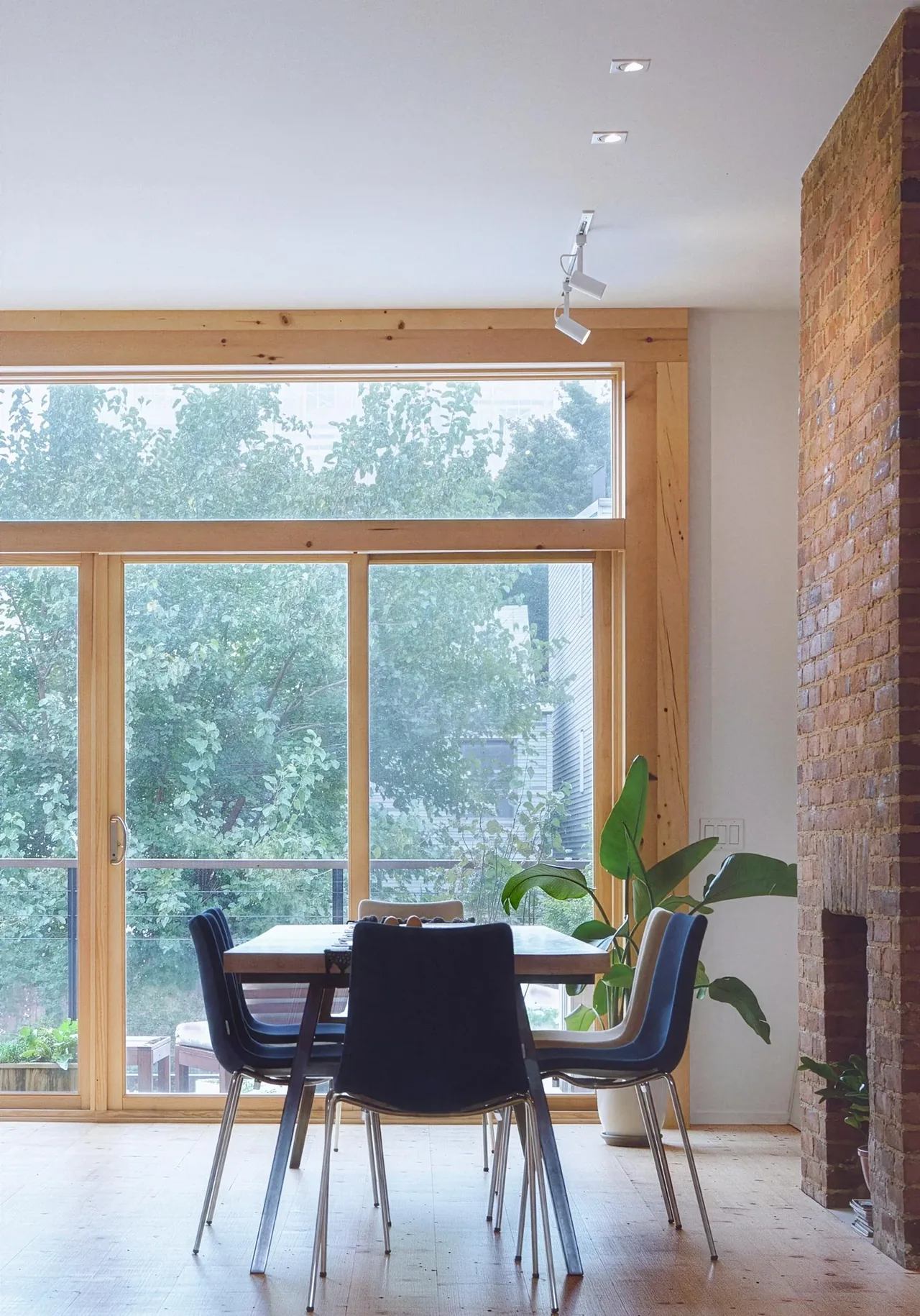 A dining space features a wood table, dark chairs, large wood-framed sliding glass doors, and an exposed brick wall in the Brooklyn Townhouse Renovation.