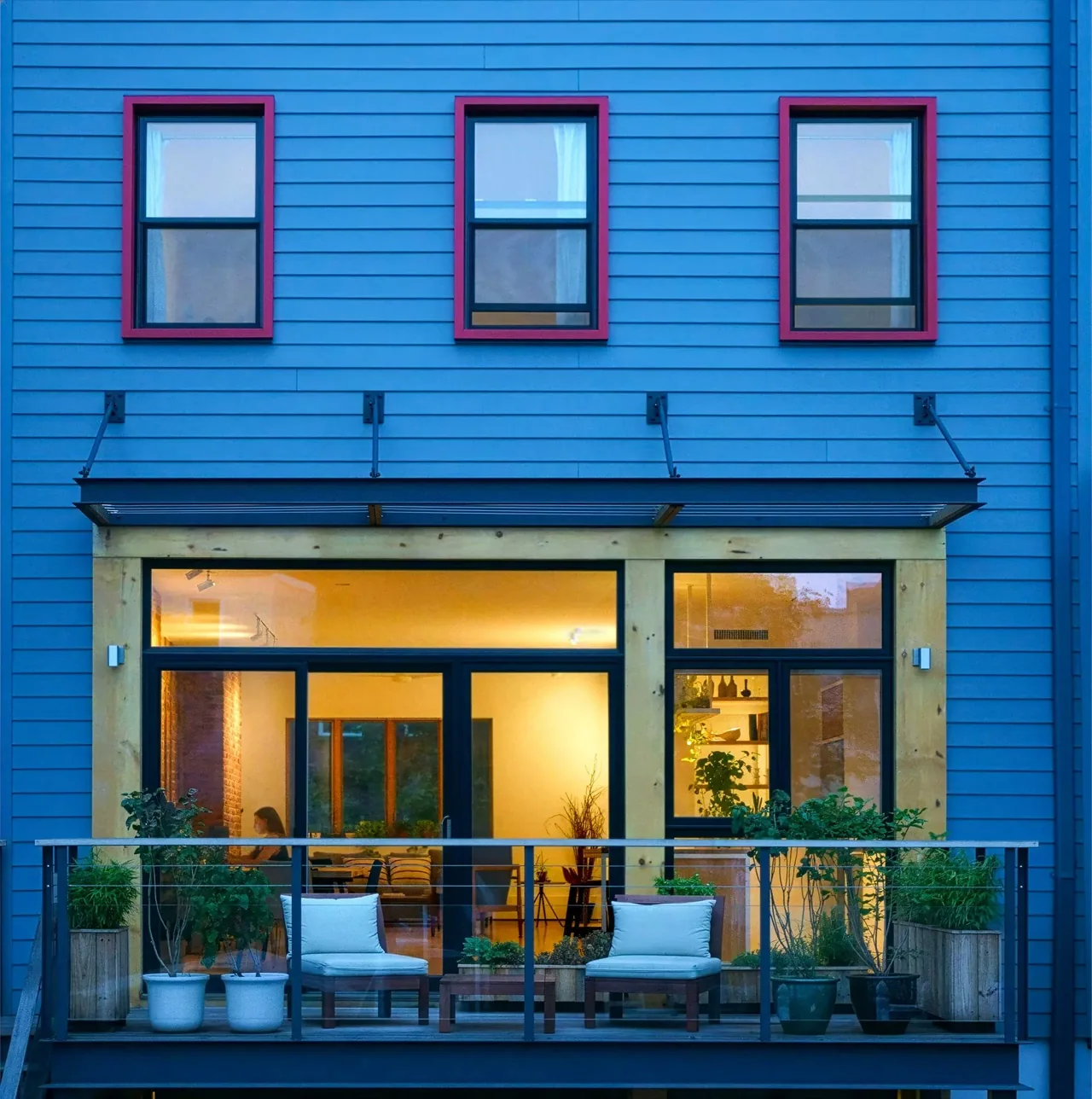 Brooklyn townhouse with blue siding, red window frames, and a balcony with seating and plants.