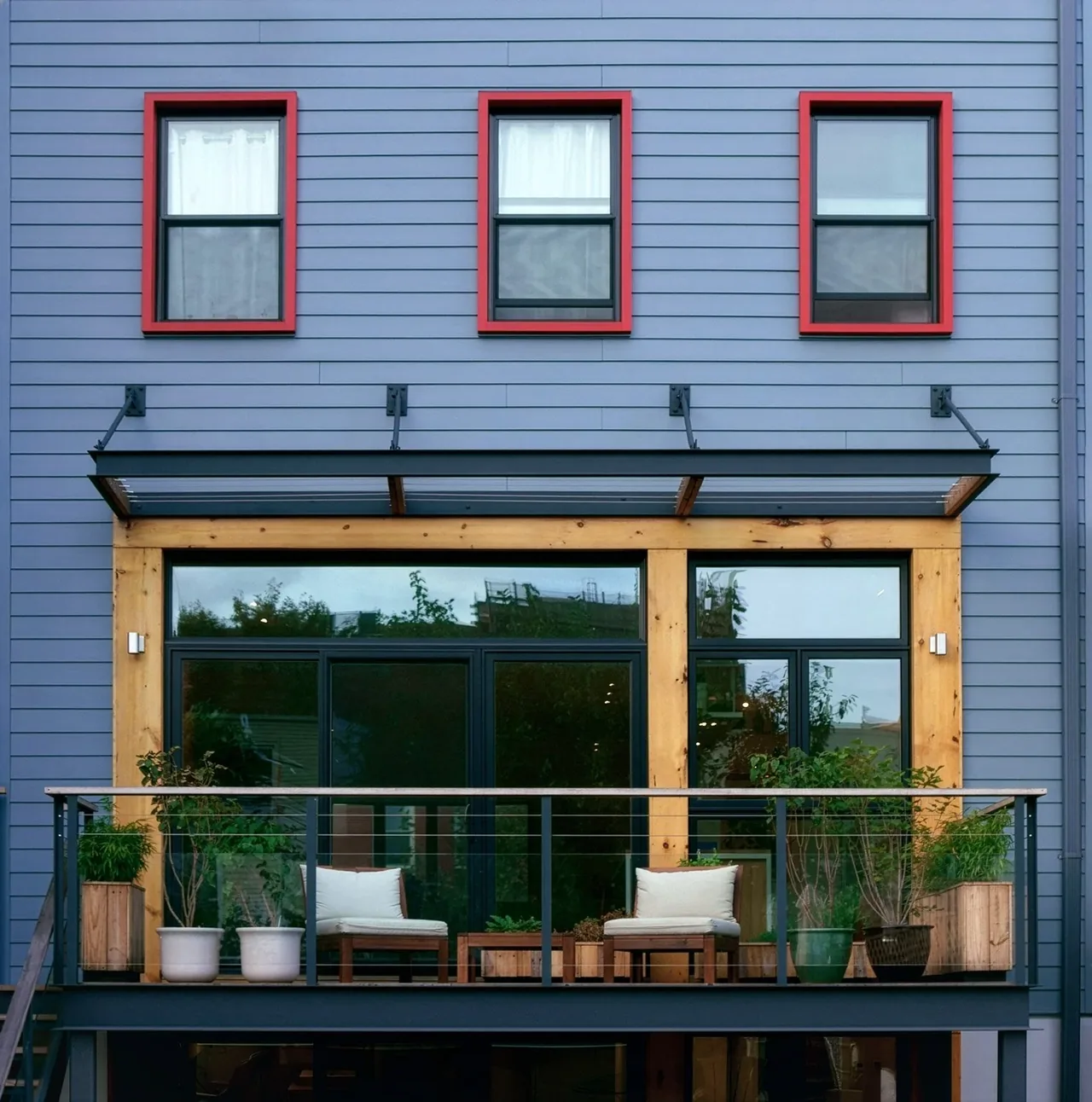 Townhouse exterior with blue siding, red window trim, black framed balcony, and a wood-trimmed large glass door opening to a patio.