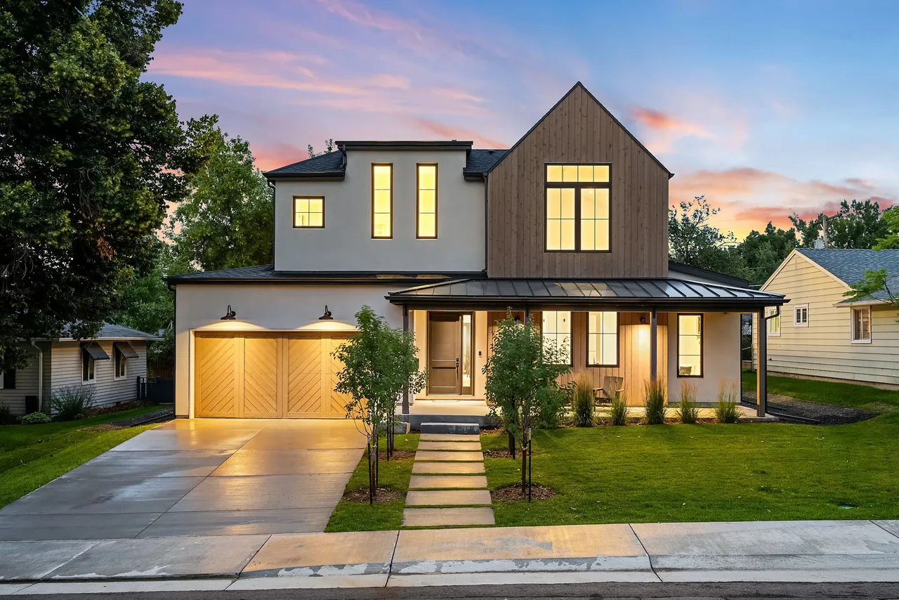 Exterior front view of a modern two-story house with mixed materials including stucco and wood siding, a covered porch with a metal roof, and multiple windows illuminated at dusk.