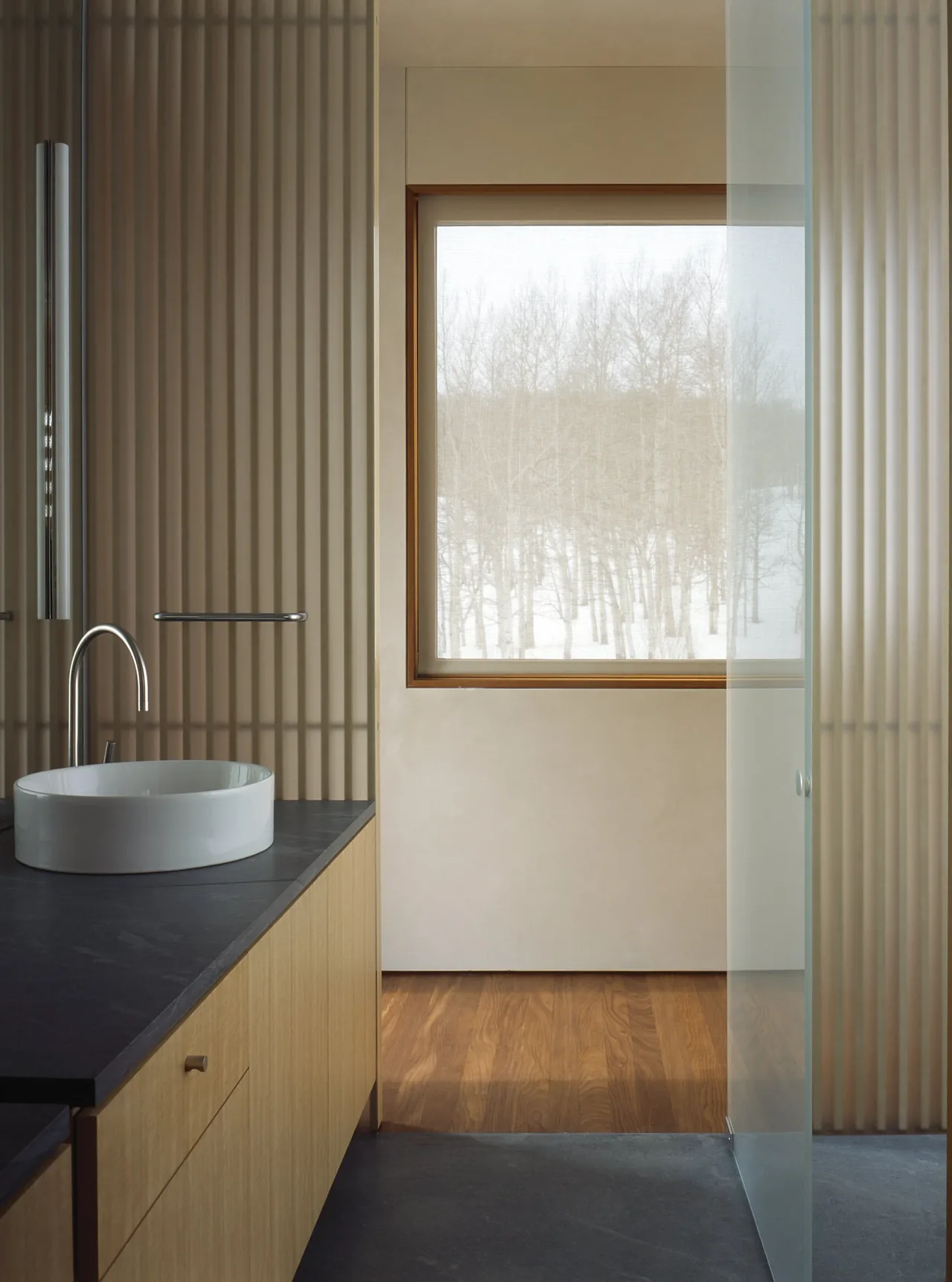 Bathroom interior with wood vanity, black countertop, vessel sink, and vertical slatted wall panels beside a window showing leafless trees outside.