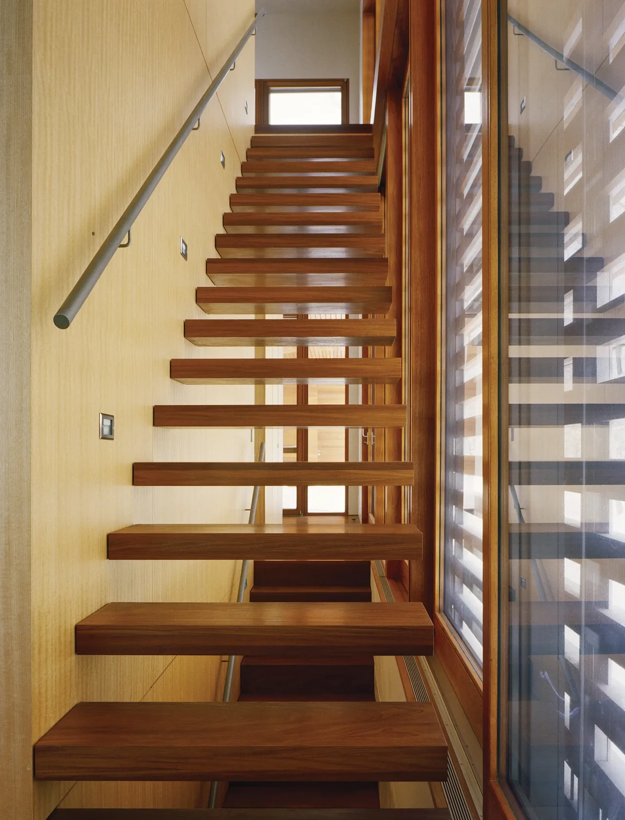 Floating wooden stairs with thick treads and a metal handrail next to a large window reflecting the stairs.