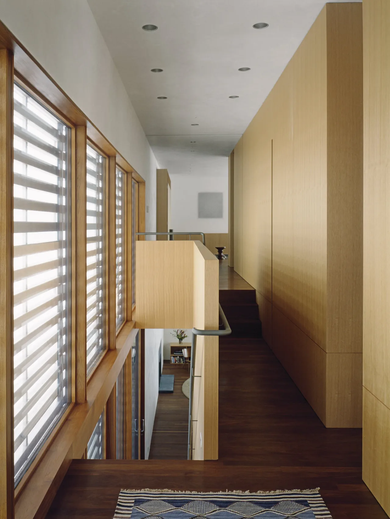 A wooden-paneled hallway with windows featuring horizontal striped shades and recessed ceiling lights.