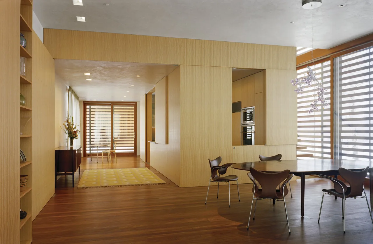 The dining area features a round table with four chairs next to a window with sliding shades, adjacent to a hallway with wood paneling and flooring.