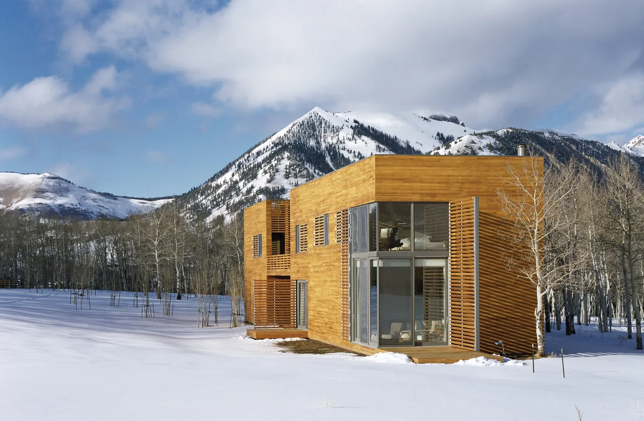 Exterior view of a wooden rectangular house featuring large floor-to-ceiling windows and wood slat screens in a snowy mountain setting.