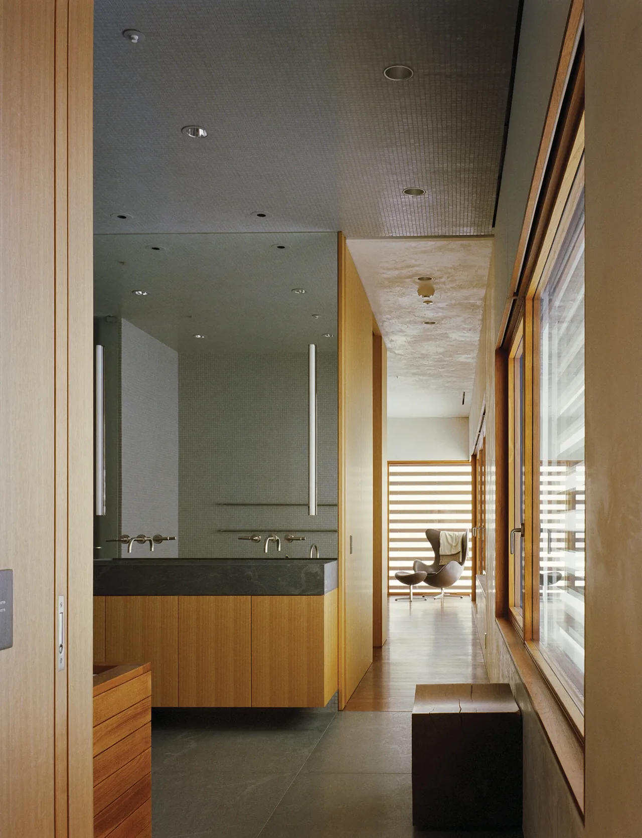 Interior view of a bathroom hallway with natural wood finishes and a seating area at the end near large windows with horizontal blinds.