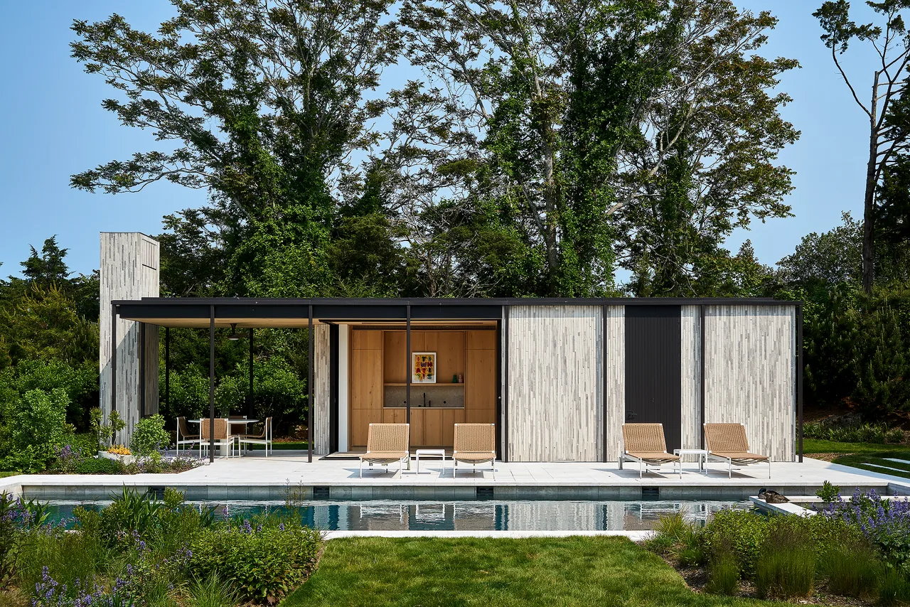 Exterior view of a modern pool house featuring vertical light wood panels, an attached pergola with a dining area, and poolside lounge chairs.
