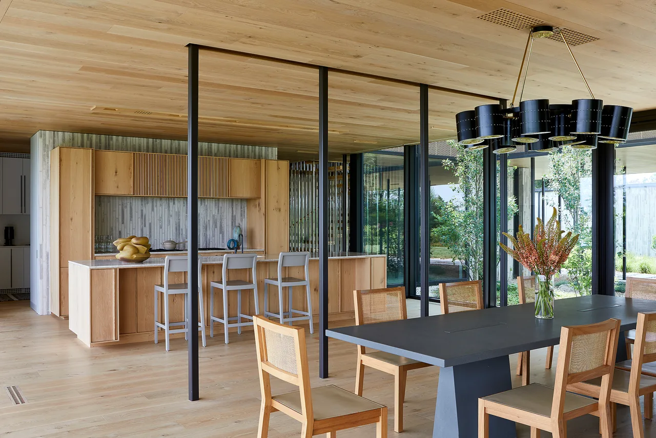 Open-plan dining and kitchen area featuring wood cabinetry, floor-to-ceiling glazing, and a contemporary chandelier.