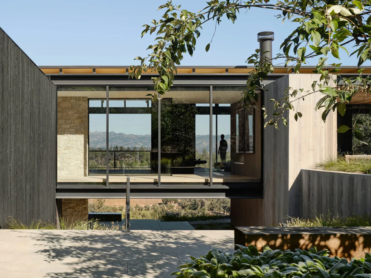 Elevated glass walkway with stone and concrete walls connects sections of the Trestle Residence, showcasing mountain views.