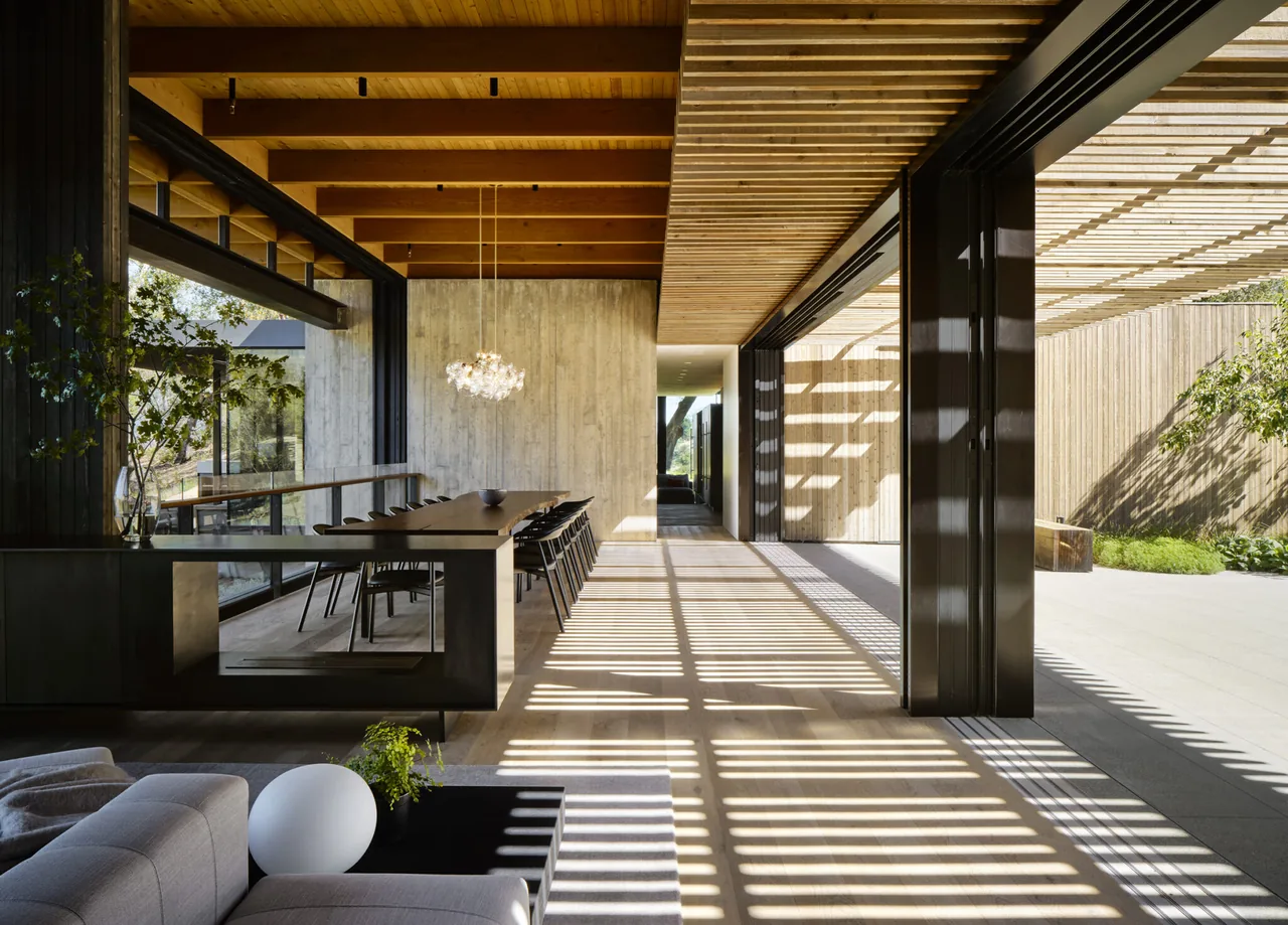 Dining space with wood ceiling beams opens to an outdoor patio covered by a wood pergola allowing striped light patterns.