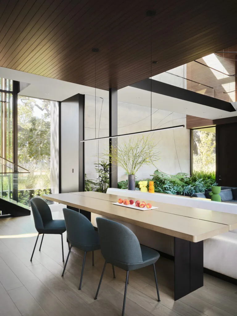 Dining area features large wood table, upholstered chairs, and built-in bench seating with views to landscaped exterior through floor-to-ceiling windows.