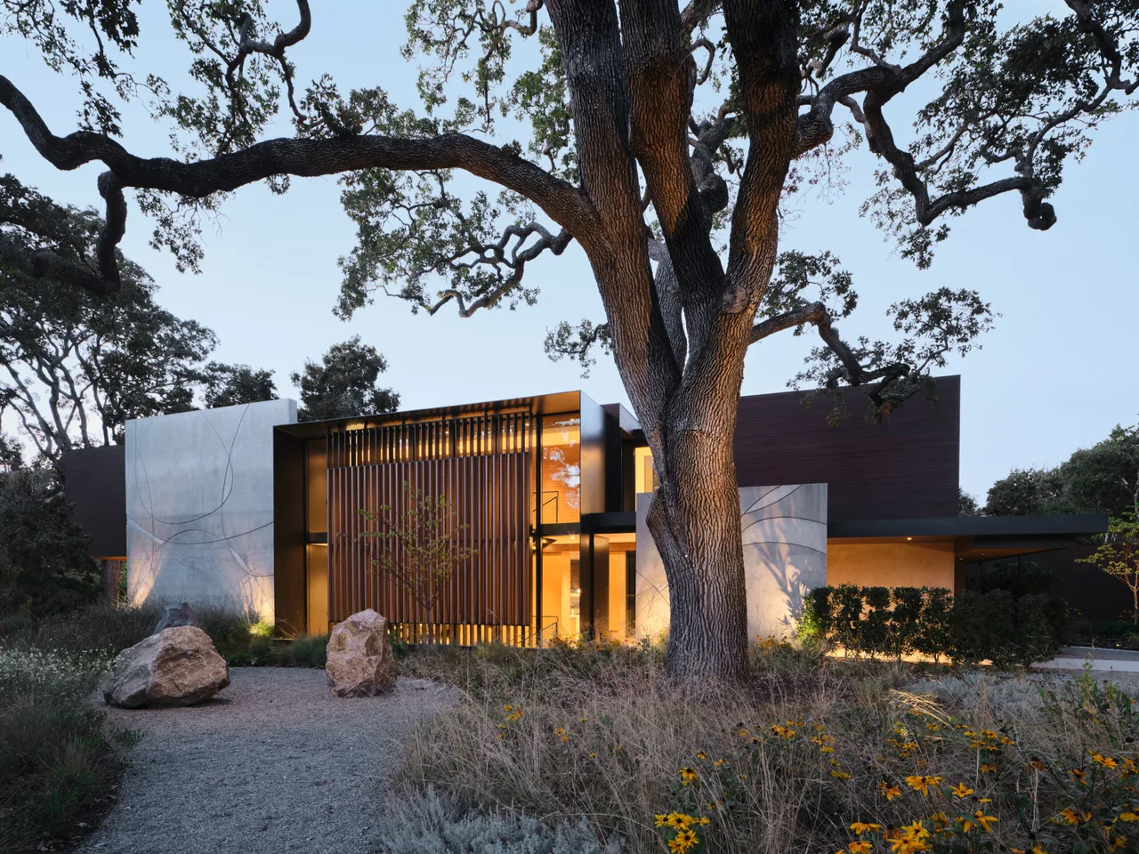 Exterior front view of a modern residence featuring concrete walls, vertical wood slats, and large windows at dusk.
