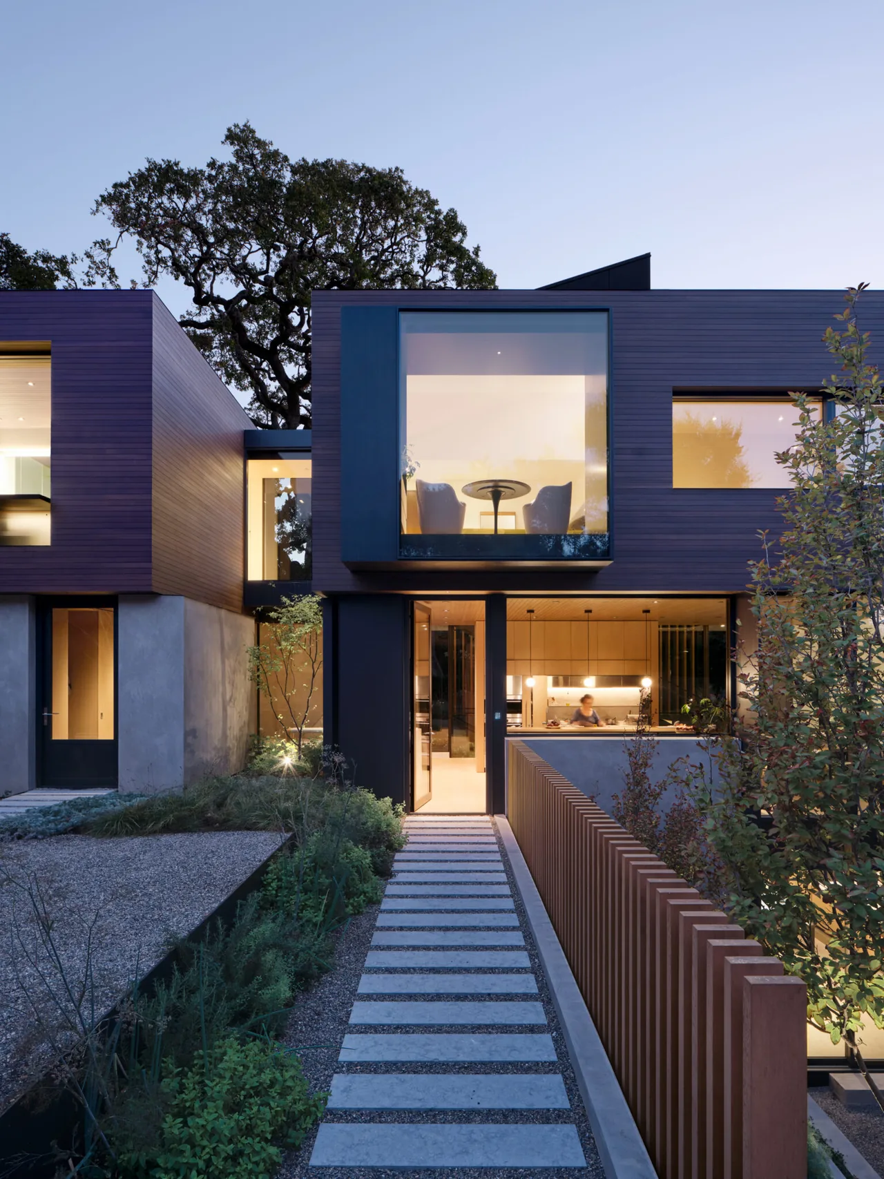 Exterior front view of modern two-story residence with large corner window and illuminated kitchen visible through glass doors at dusk.
