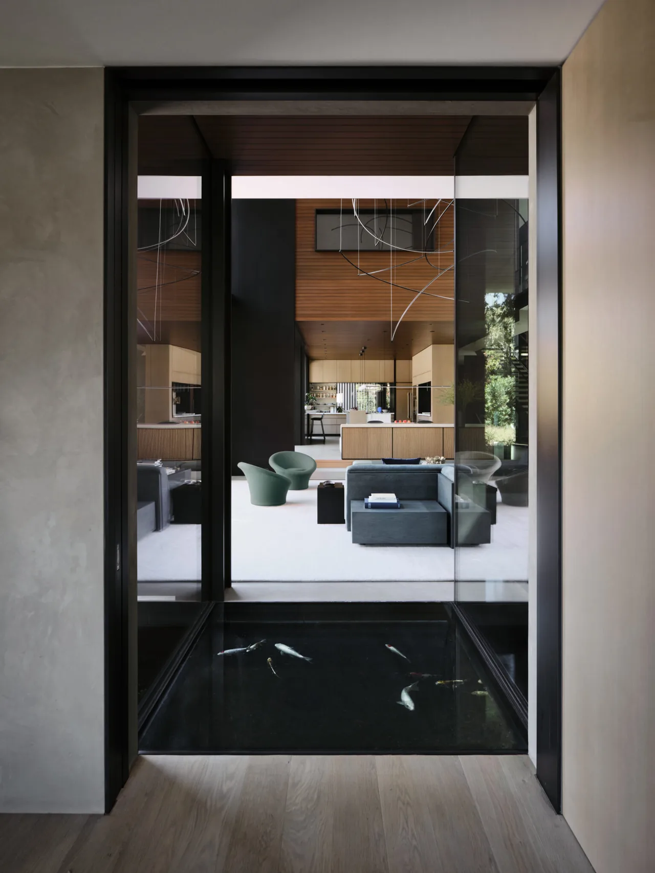 Interior view from a hallway through a glass-enclosed koi pond into a living room with modern furnishings and wood ceiling.