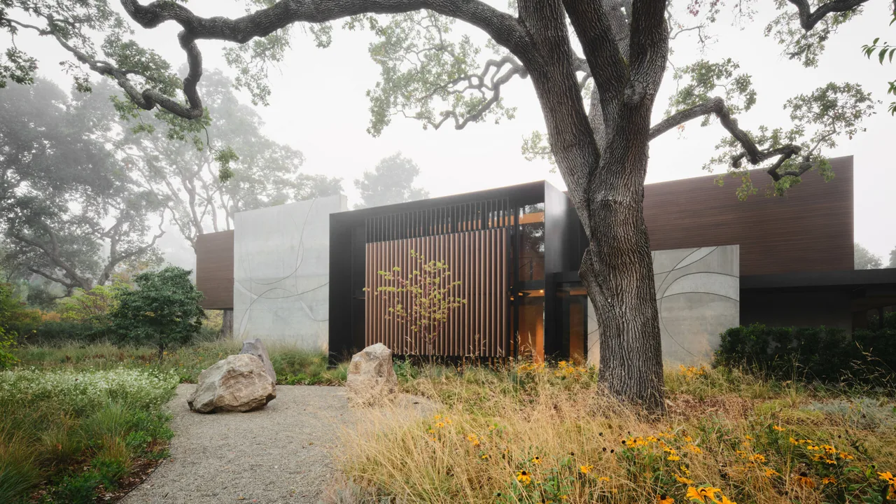 Exterior front view of a modern residence with wood siding and decorative concrete panels surrounded by natural landscaping.