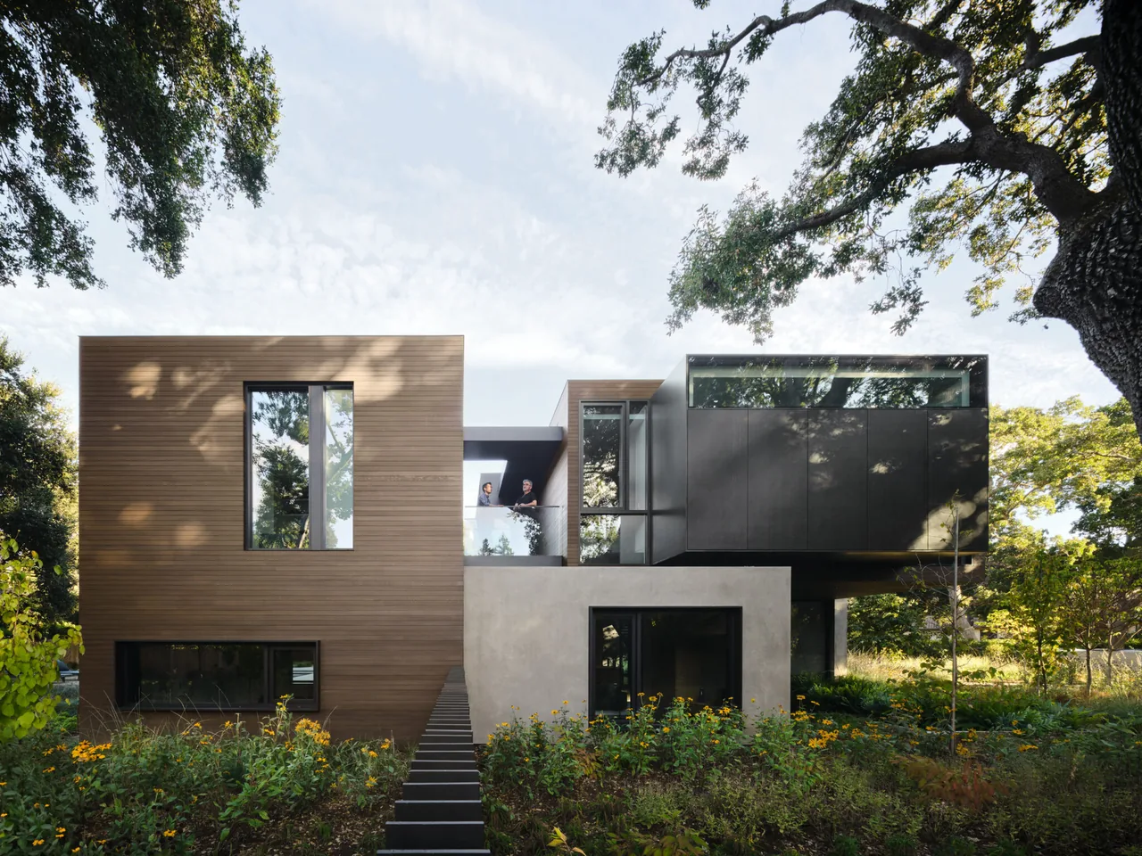 Exterior front view of a modern cubic residence clad in wood, dark metal panels, and concrete with large windows and landscaping.