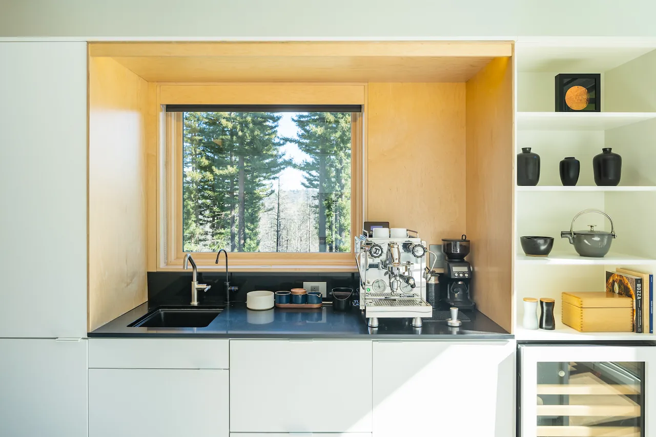 Kitchen counter with integrated sink and espresso machine beneath a large window framed by light wood.