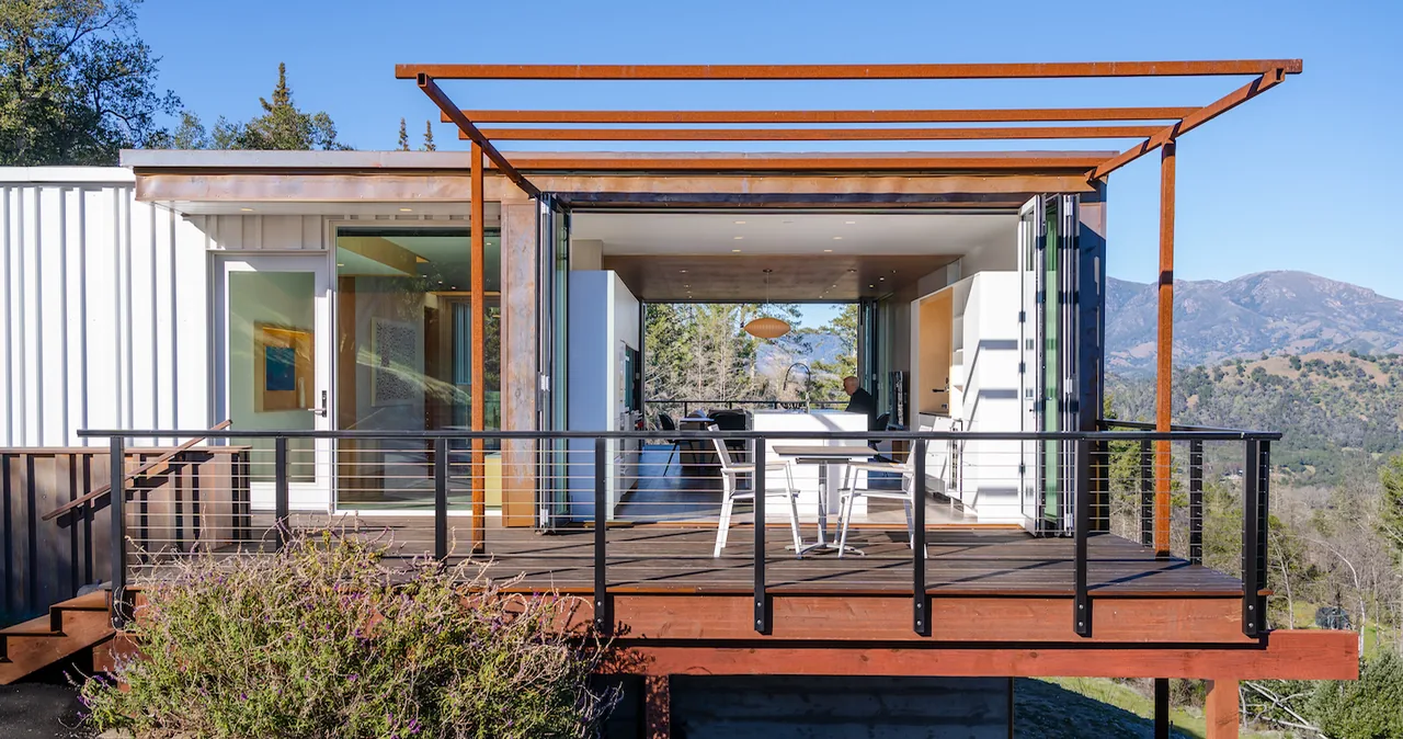 Rear exterior view showing a wooden deck with outdoor seating, metal railing, and folding glass doors connecting to the kitchen interior.
