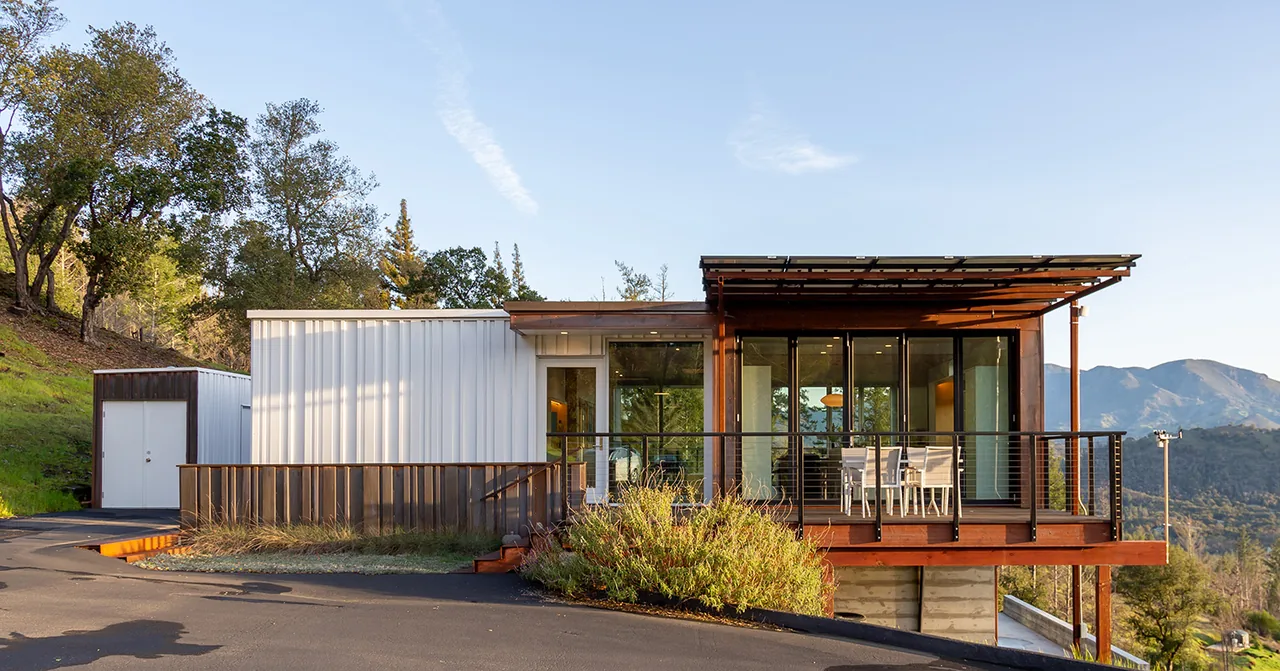 Front exterior of a modern house featuring white metal siding, a wooden deck, and large glass doors opening to the deck.