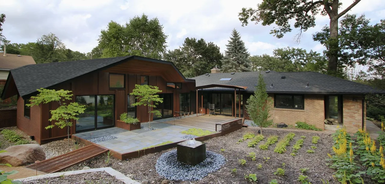 Folded House exhibits a blend of wood siding and brick with large windows and an outdoor stone patio in a landscaped backyard.