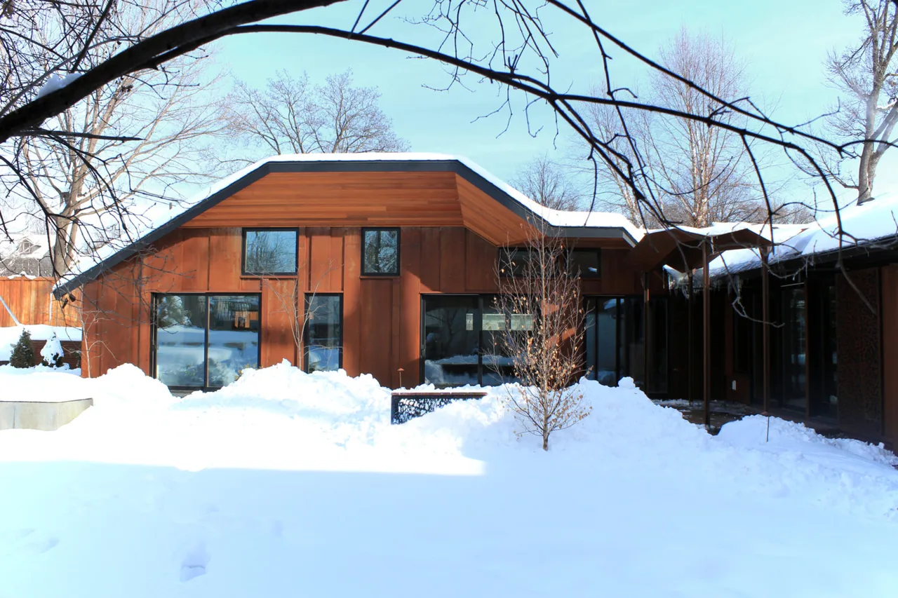 Winter exterior rear of the Folded House showing weathered metal siding, wood soffit, and black-framed windows.