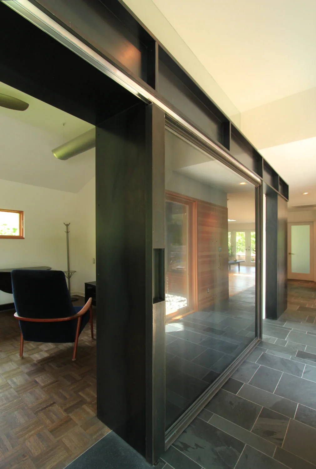 Sliding glass door with a black metal frame separates a tiled corridor and a room with wooden parquet flooring.