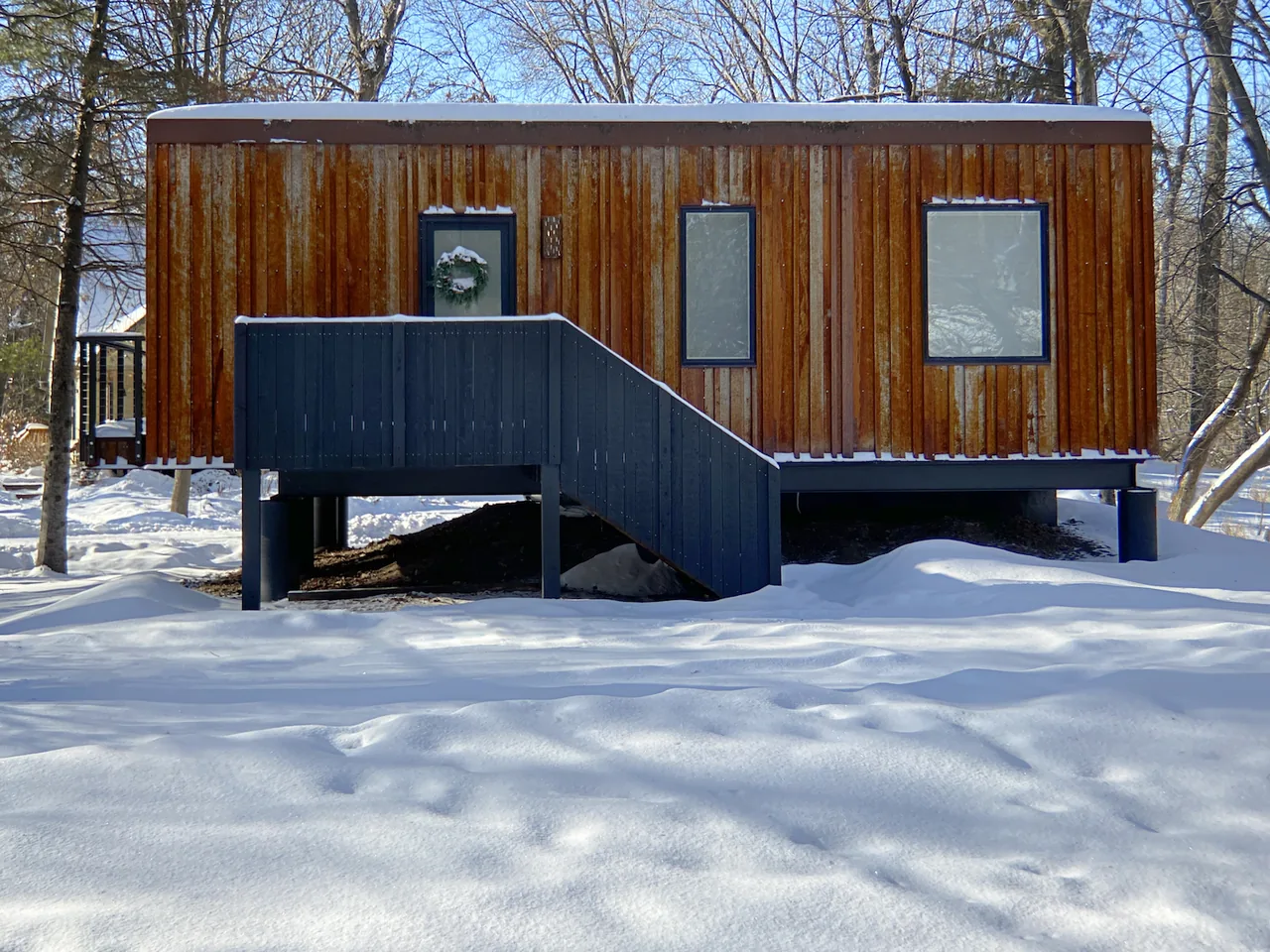 Elevated house with rusted vertical metal siding and black wooden staircase in snowy landscape.
