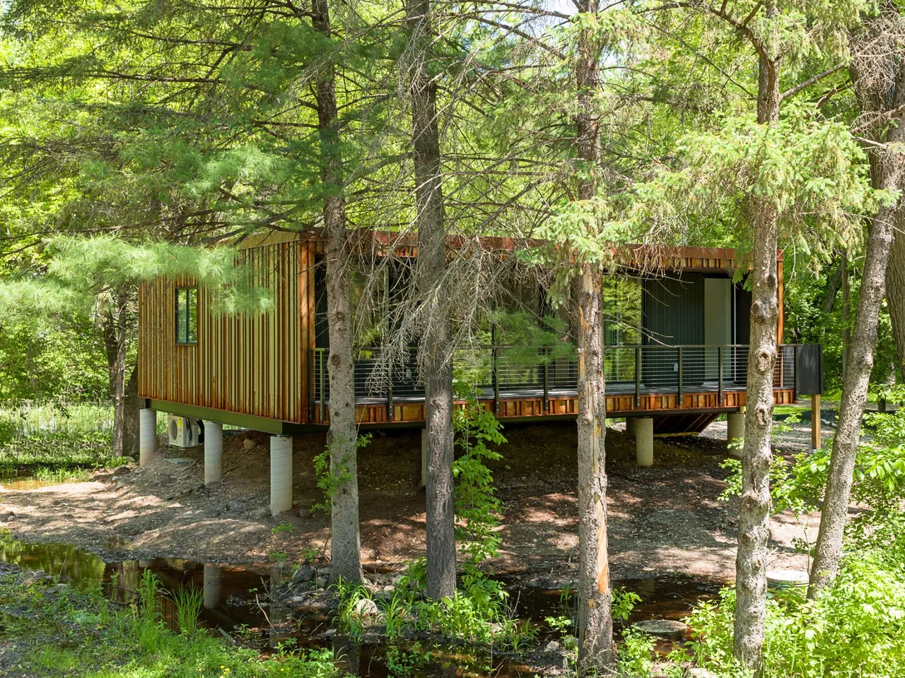 The weeHouse is a compact elevated structure with vertical wood cladding and floor-to-ceiling windows set in a wooded, natural environment.