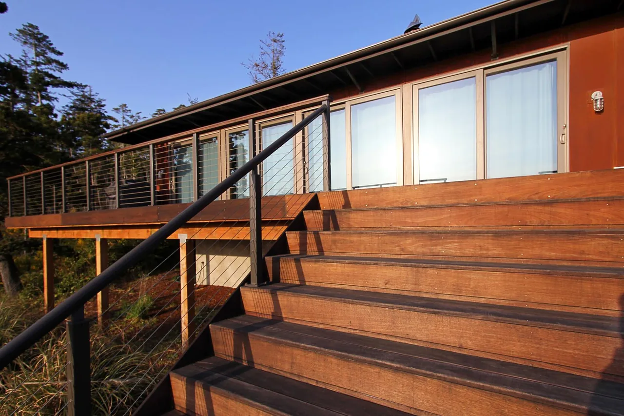 Exterior front of Oceanside weeHouse features wide wooden stairs with cable railings leading to a deck with floor-to-ceiling glazing windows.