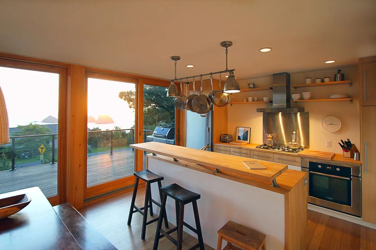 Oceanside weeHouse kitchen with wood finishes and stainless steel appliances opens to a deck with glass railings and ocean views.