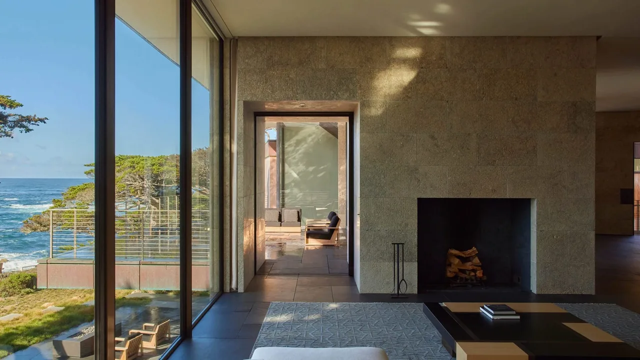 The Dogleg Residence living room has a large fireplace and floor-to-ceiling windows framing a waterfront view in sunset lighting.