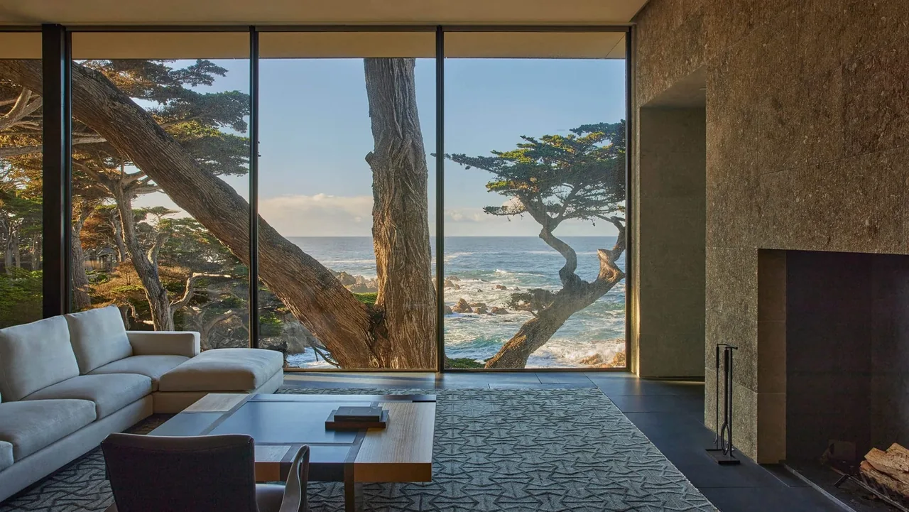 Living room in Dogleg Residence with floor-to-ceiling glazing and stone fireplace overlooking waterfront vegetation and ocean.