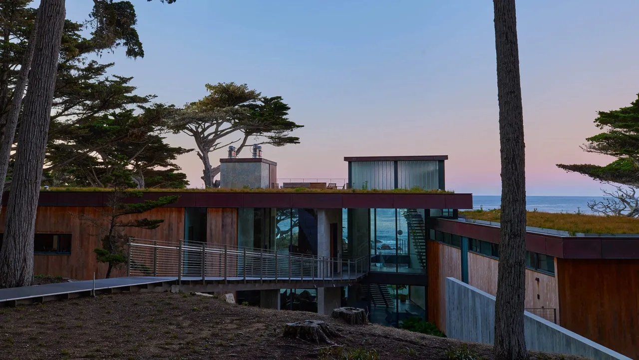 The Dogleg Residence features a green roof, glass walls, and wood siding with ocean views, blending into the natural landscape.