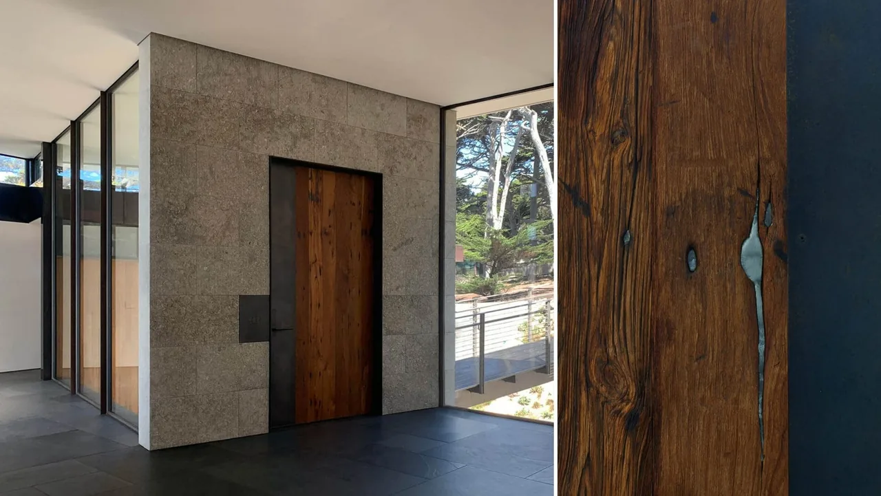 Interior corridor featuring a wooden door inset in a stone wall, slate tile flooring, and black-framed floor-to-ceiling windows revealing an outdoor walkway.