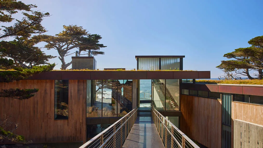 Modern Dogleg Residence features wood siding, extensive glazing, and a flat green roof with ocean views.