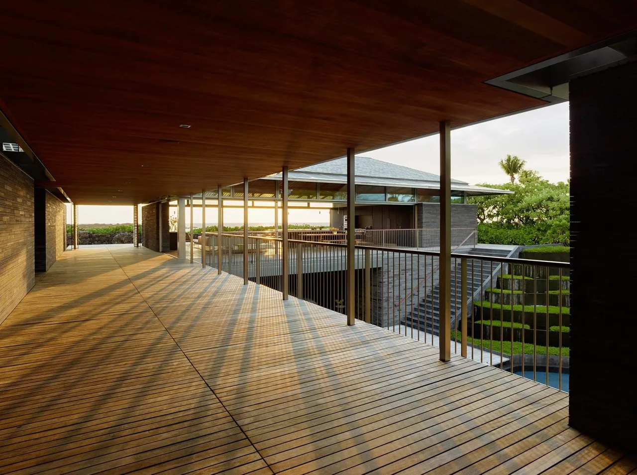 Covered wooden deck corridor with metal railings and views of landscaped garden and pool area.