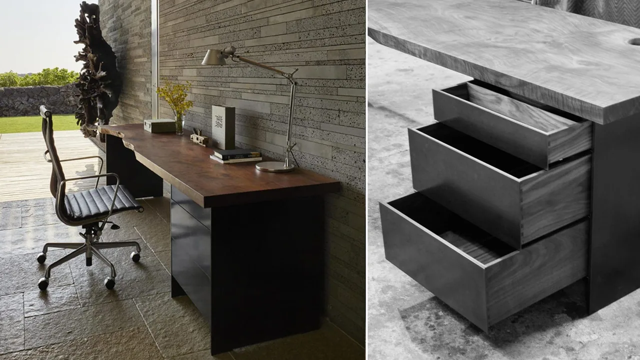 Work desk with wooden top and drawers beside a textured stone-tiled floor and wall overlooking an outdoor grassy area.
