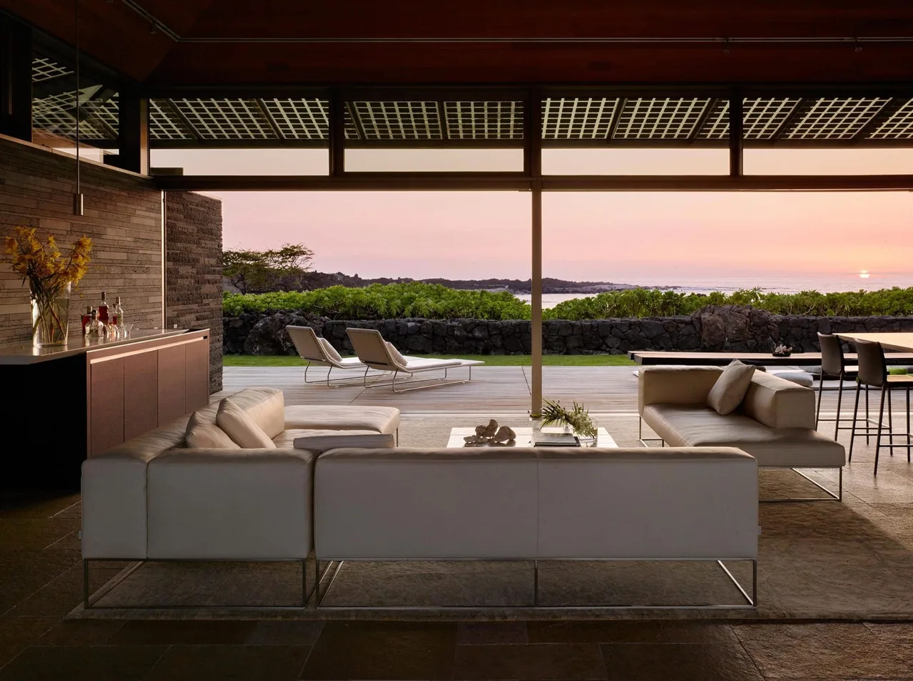 Living room opens via sliding glass doors to a wooden deck overlooking an ocean at sunset.