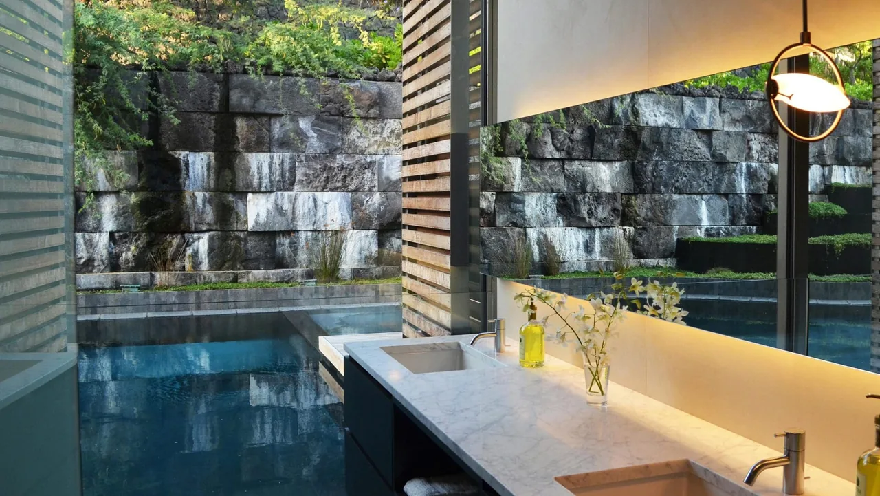 Bathroom with marble double sink counter adjacent to a pool and stone wall seen through large glass windows.