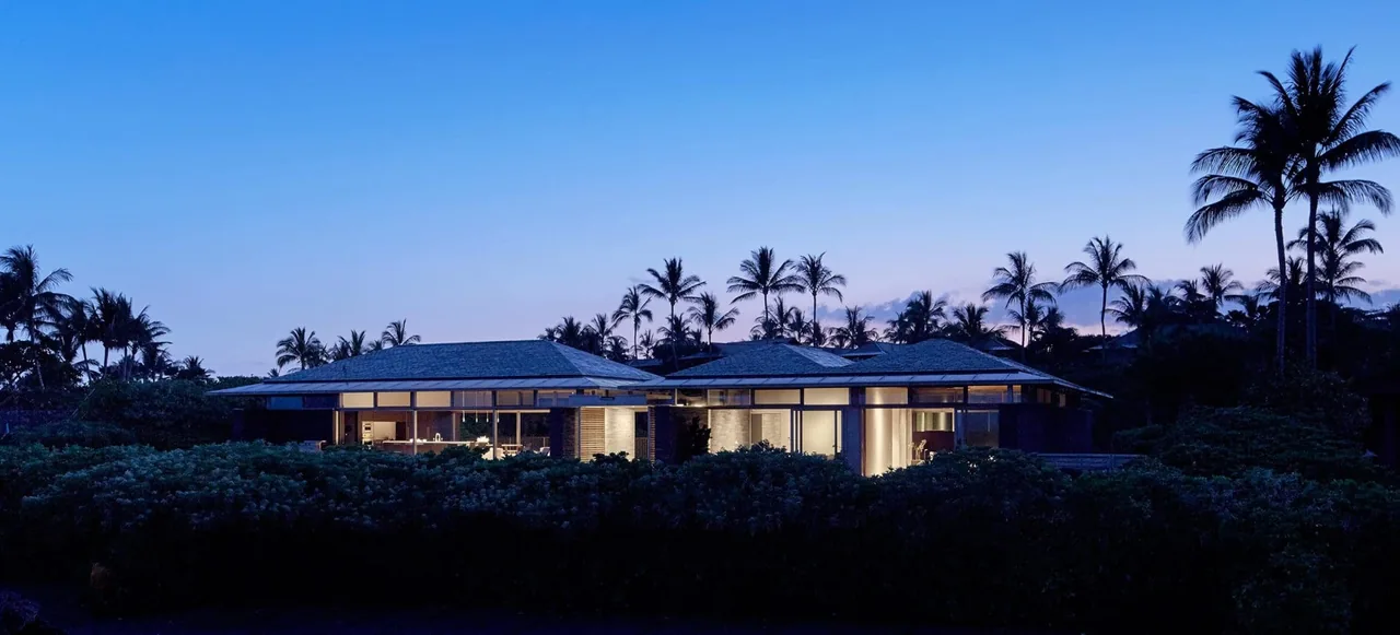 Wide exterior front view of a modern residence at dusk showing expansive glass walls and hipped roofs.