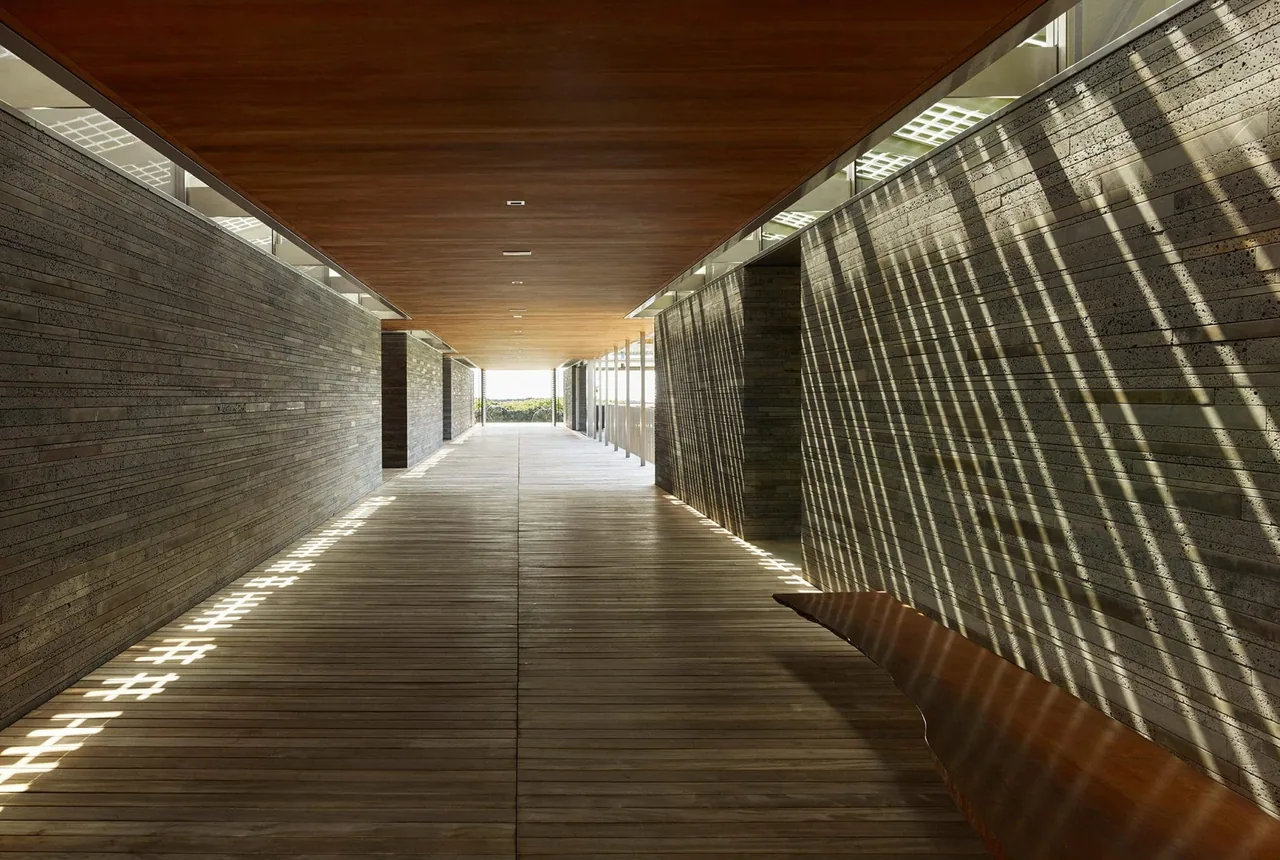 Interior corridor with wood floors and ceiling, and textured horizontal concrete walls casting shadows through slatted openings.