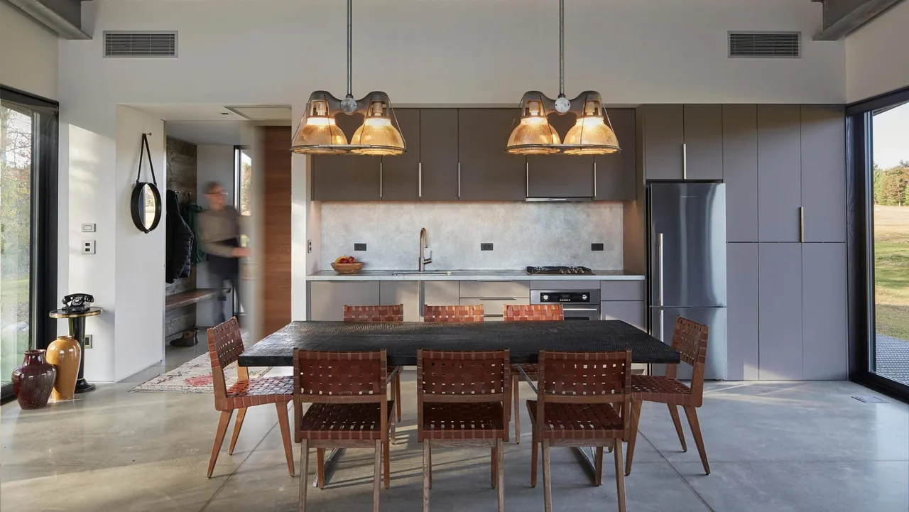 The Flying Dollar Residence kitchen features gray cabinetry, welded leather dining chairs, and pendant lighting with large windows overlooking an autumn landscape.