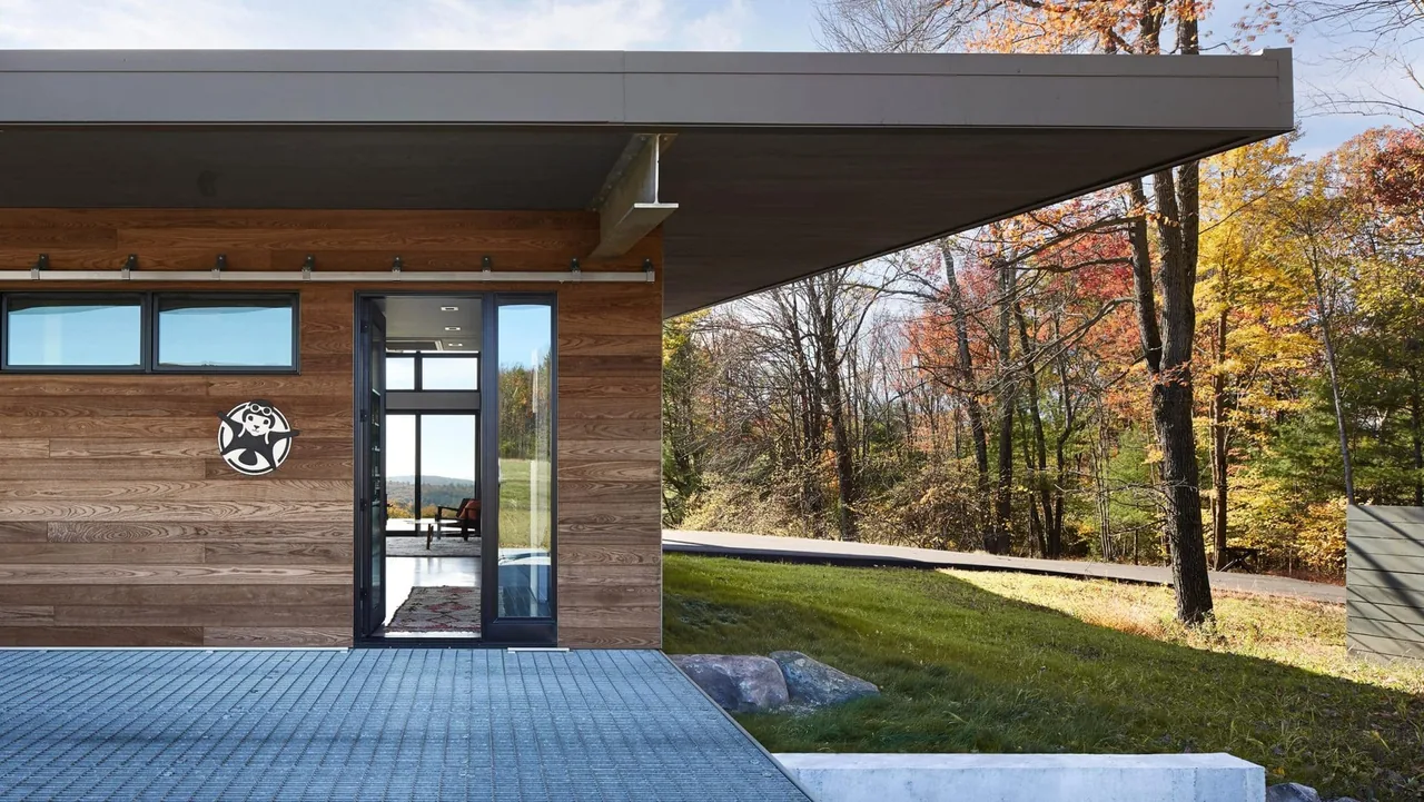 The Flying Dollar Residence features horizontal wood siding, floor-to-ceiling glazing in the entry door, and a flat roof with a cantilevered overhang.