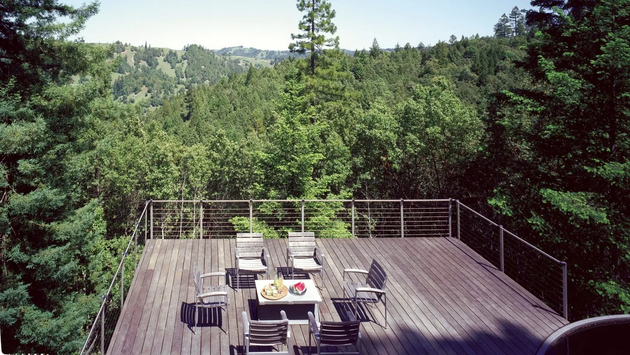 Outdoor wood deck with metal cable railing furnished with seating and a table, overlooking a wooded hillside.