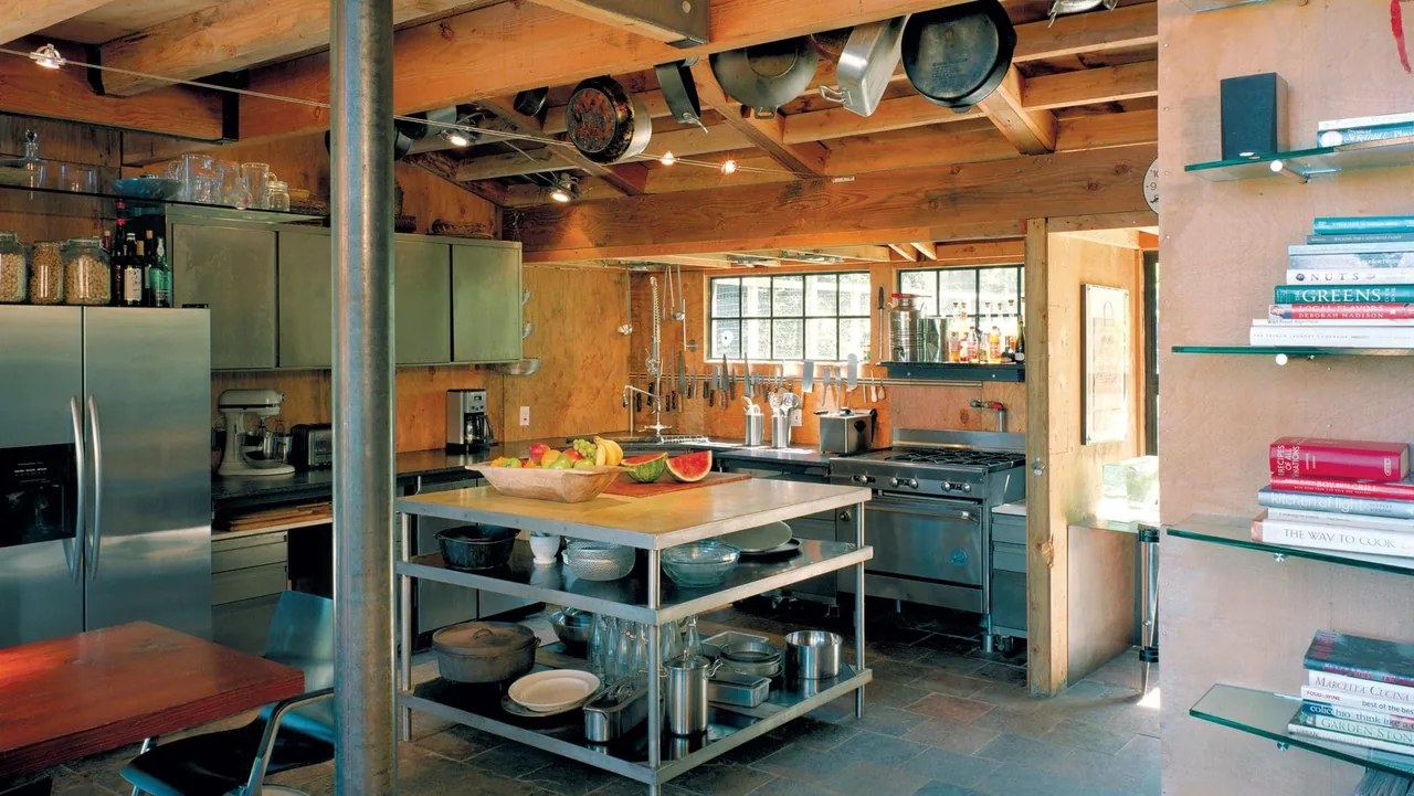 The kitchen in the Lundberg/Breuer Cabin features exposed timber beams, stainless steel appliances, and open metal shelving.