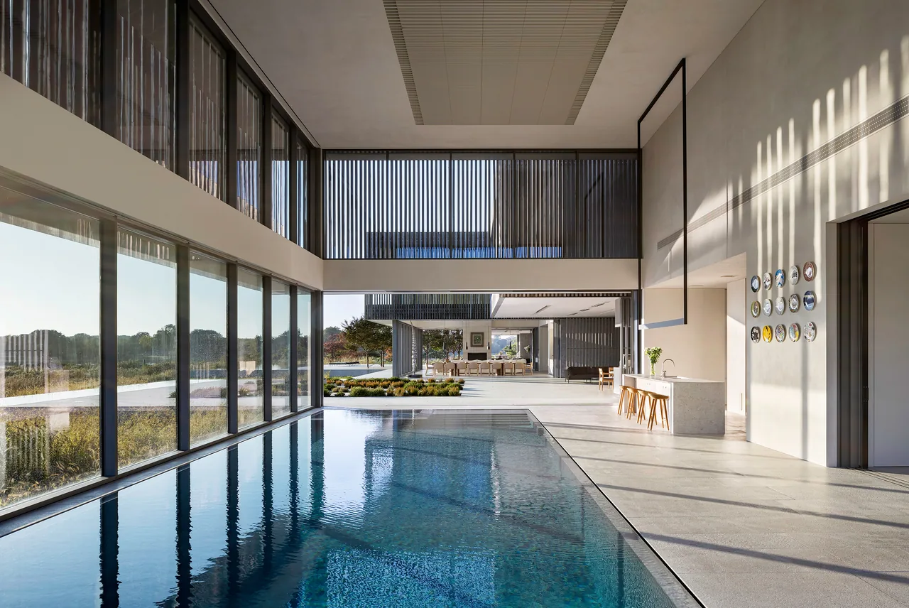 Interior view of a residential indoor pool area featuring expansive glazing and an open connection to an adjacent kitchen and outdoor patio.