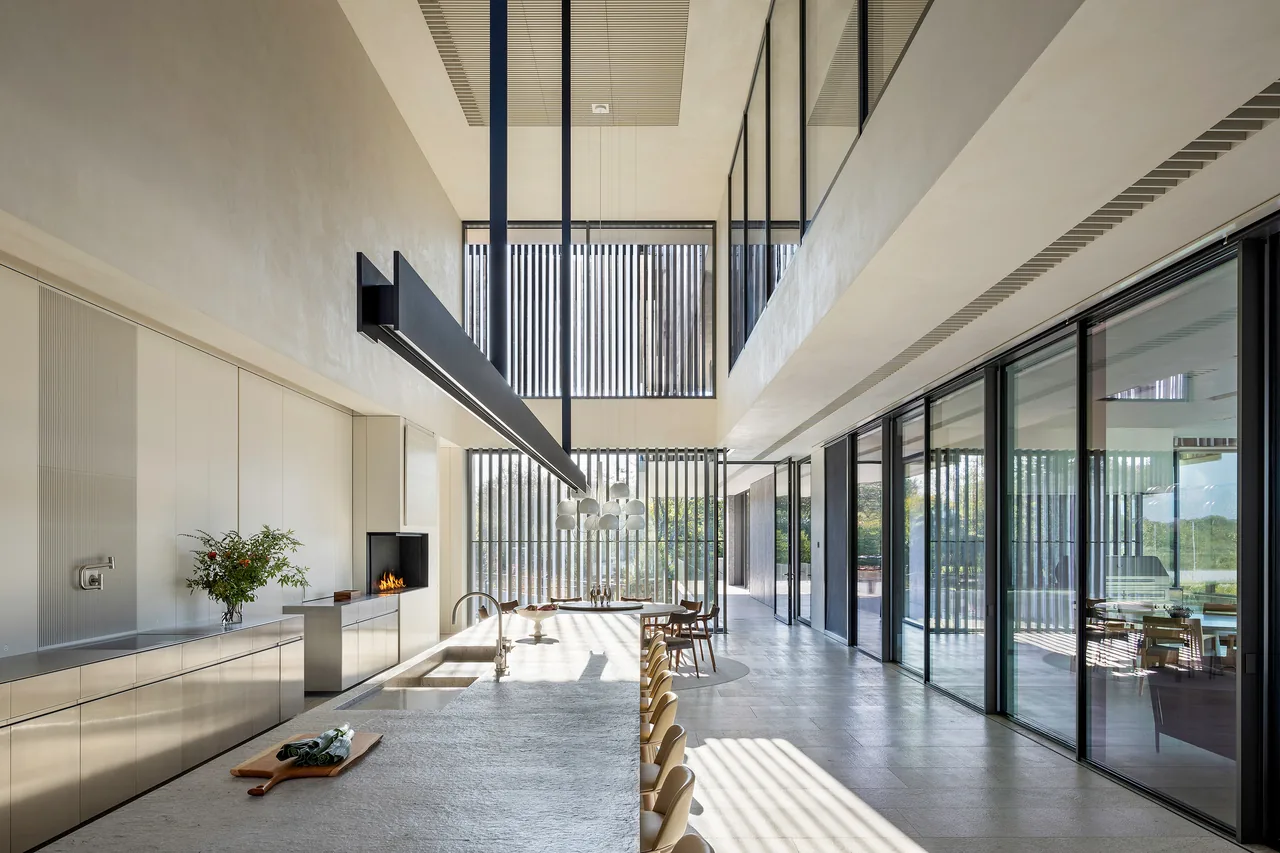Modern kitchen and dining area with floor-to-ceiling glass doors, vertical metal louvers, and a large kitchen island under a suspended linear light fixture.