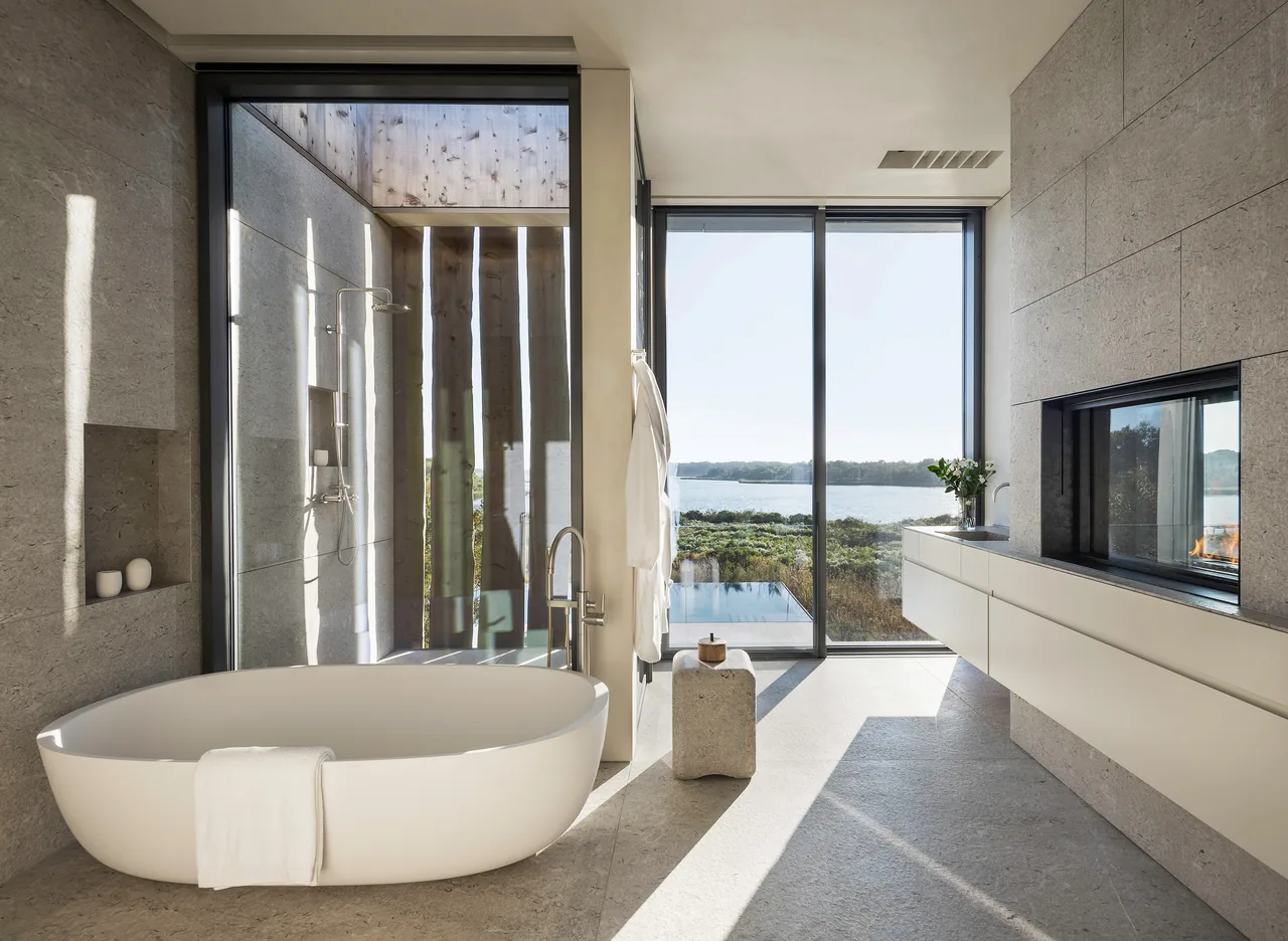 A spacious modern bathroom featuring a freestanding tub, floor-to-ceiling glass windows overlooking a water view, and a built-in fireplace.