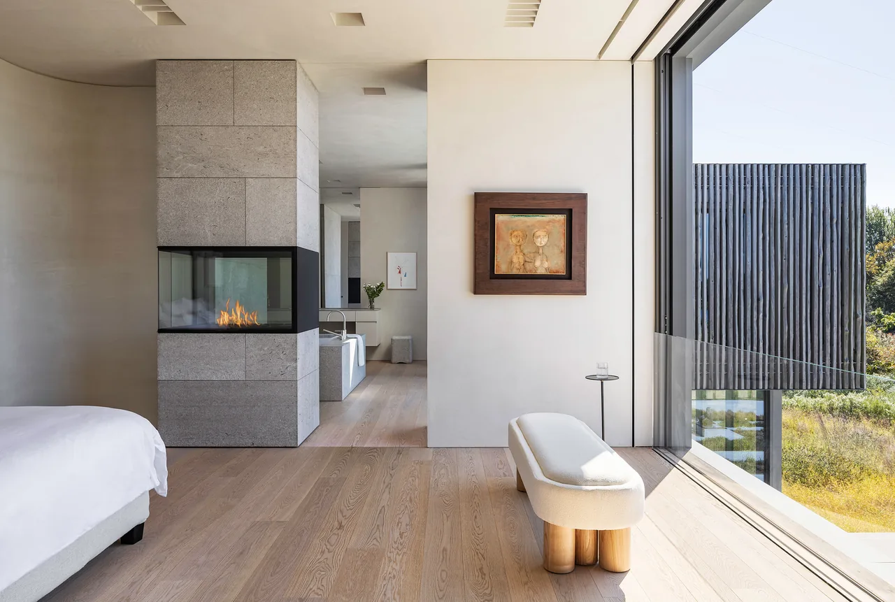 Bedroom featuring a three-sided glass fireplace set in a stone column and large floor-to-ceiling window with a view of exterior vertical wood elements.